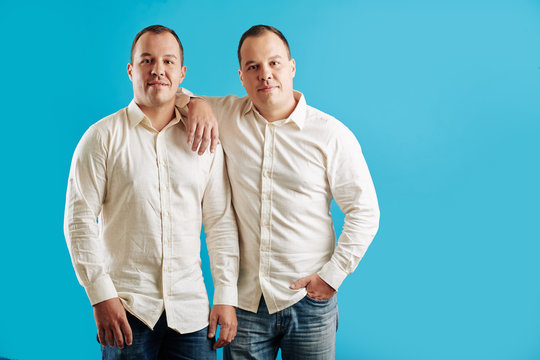 Two Identical Male Twins Wearing White Shirts And Jeans Standing Against Blue Background Looking At Camera