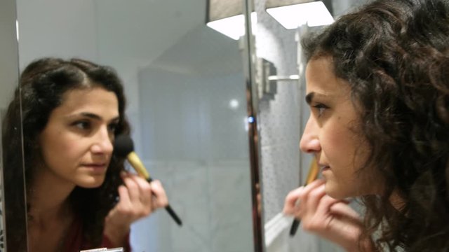 A beautiful middle eastern brunette woman preparing for date applying makeup powder  on the cheekbones while looking sad in mirror. 4k