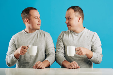 Young adult male twin siblings sitting at table looking at each other holding cups with coffee,...