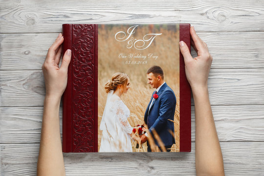 Red wedding photo book with leather cover. stylish wedding photo album close up. person opens a square photobook. family burgundy photoalbum on wooden background. womans hand holding a  photo album