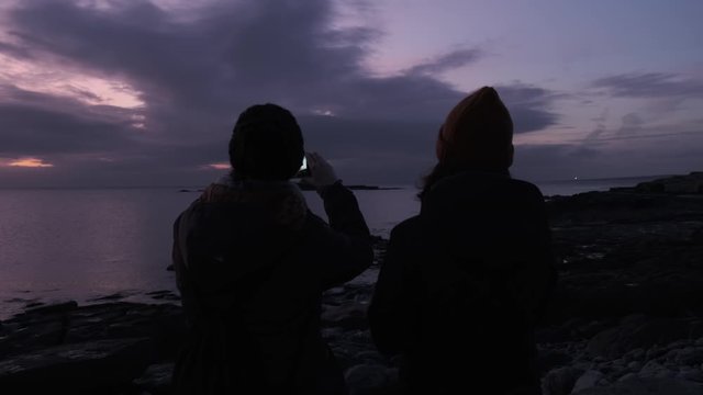 Two women looking at the sunset in Ireland and talking a photo with a smartphone. Beautiful cloud formations.