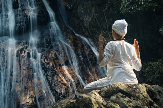 Young adult woman in white clothes sits on a rock against the background of a waterfall and practices yoga
