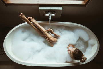 Woman relaxing in foam bath with bubbles in dark bathroom by window
