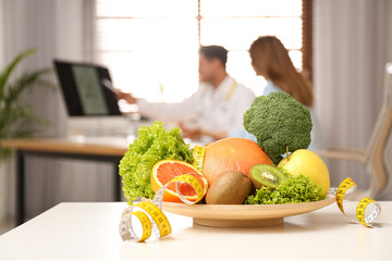 Nutritionist consulting patient at table in clinic, focus on plate with fruits, vegetables and...