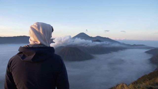 Young man with hoodie and outdoor jacket overlooking volcanic landscape of Bromo Tengger Semeru Nationalpark with active volcano and Sea of Sand covered in fog in early morning light from viewpoint 