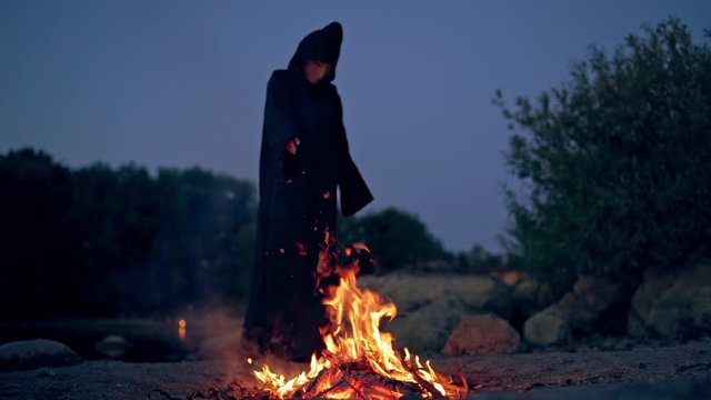 Witch evil in black gown near fire at night. Mysterious wizard doing shamanism over the flame. Scary figure in black making ritual. Halloween night.