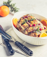 Roasted pork in white dish, christmas baked ham with cranberries, tangerines, thyme, rosemary, garlic on light table surface.