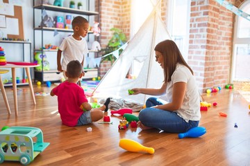 Young beautiful teacher and toddlers playing around lots of toys at kindergarten