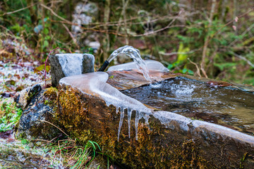 Brunnen im Winter mitten in Gebirgsschlucht, Holztrog der eine Quelle fasst mit vereisten...