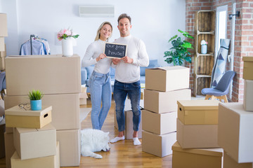 Young beautiful couple with dog standing holding blackboard with message at new home around cardboard boxes