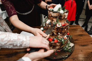  Elegant wedding cake decorated with roses and fruits with bride and groom on background. Wedding ceremony. Beautiful pie on the table. Brides cut cake together. Close up