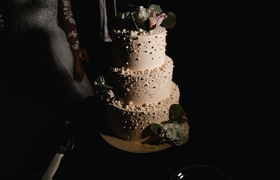  Elegant wedding cake decorated with white pearls, flowers and vanilla glaze with bride and groom on background. Wedding ceremony. Beautiful pie on the table. Brides cut cake together. Close up