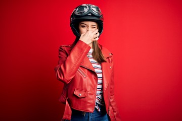 Young beautiful brunette motocyclist woman wearing motorcycle helmet and red jacket smelling...
