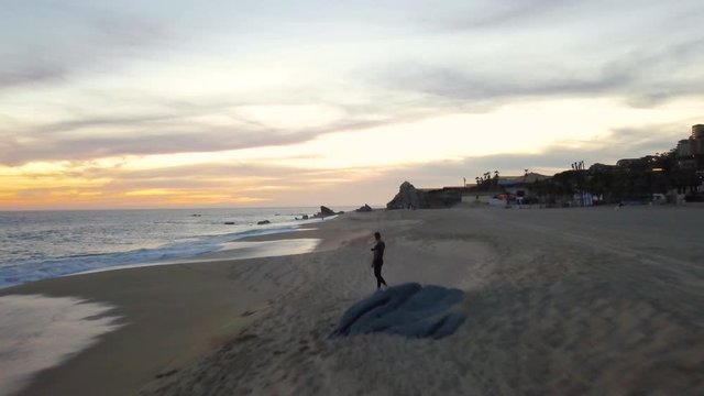 Young male standing on the shore of a beach in Cabo, San Lucas, Mexico, at sunset. Aerial flyover