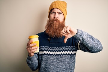 Redhead Irish man with beard wearing winter hat drinking take away hot coffee with angry face,...