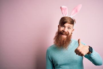 Hipster Irish man with beard wearing easter rabbit ears over isolated pink background doing happy thumbs up gesture with hand. Approving expression looking at the camera showing success.