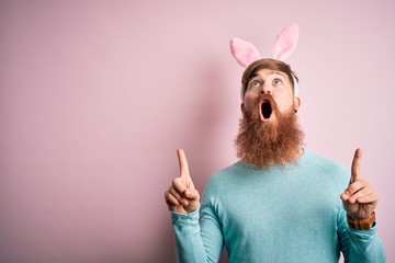 Hipster Irish man with beard wearing easter rabbit ears over isolated pink background amazed and surprised looking up and pointing with fingers and raised arms.