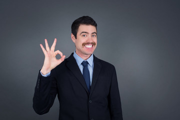 Glad attractive man shows ok sign with hand as expresses approval, has cheerful expression. Photo of handsome male has appealing appearance, being optimistic. Standing against gray wall.