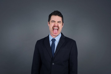 Beautiful caucasian man with happy and funny face smiling and showing tongue. Wearing casual clothes and standing against gray studio background.