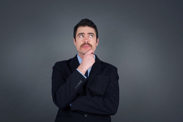 Portrait of thoughtful  man keeps hand under chin, looks away trying to remember something or listens something with interest, dressed casually, poses indoors. Youth concept.