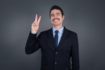 Young caucasian man standing against gray wall showing and pointing up with fingers number two while smiling confident and happy.