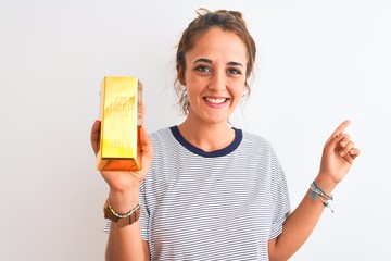 Young beautiful redhead woman holding gold ingot over isolated background very happy pointing with...