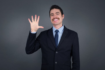 Young businessman standing against gray wall showing and pointing up with fingers number five while smiling confident and happy.