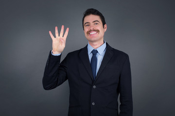 Young businessman standing against gray wall showing and pointing up with fingers number four while smiling confident and happy.