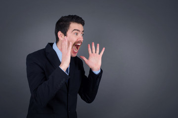 Portrait of young businessman with shocked facial expression, holding hands near face, screaming and looking sideways at something amazing. Stands against gray studio Wall.