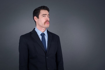 Beautiful businessman with snobbish expression curving lips and raising eyebrows, looking with doubtful and skeptical expression, suspect and doubt. Standing indoors over gray background.