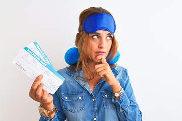 Young redhead tourist woman wearing sleeping mask and neck pillow showing boarding pass serious face thinking about question, very confused idea