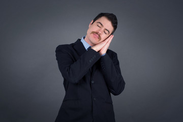 Relax and sleep time. Tired young handsome man with closed eyes leaning on palms as pillow pretending sleeping being exhausted seeing dreams standing against gray background. Sleeping gesture.
