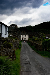 A small cottage and other farm buildings are positioned at the end of a narrow country lane with dark stormy clouds imposing above. 