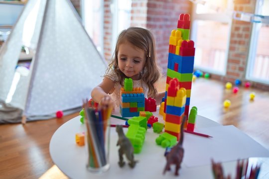 Adorable blonde toddler playing with building blocks around lots of toys at kindergarten