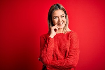 Young beautiful blonde woman wearing casual sweater over red isolated background looking confident at the camera smiling with crossed arms and hand raised on chin. Thinking positive.