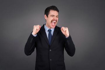 Waist up portrait of strong successful determined young businessman raising arms, clenching fists, exclaiming with joy and excitement. Victory, success and achievement concept.