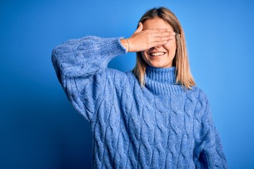 Young beautiful blonde woman wearing winter wool sweater over blue isolated background smiling and laughing with hand on face covering eyes for surprise. Blind concept.