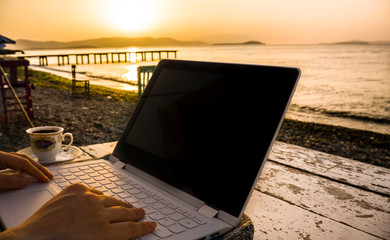 Freelance female hands working with laptop on wooden table on beach at sunset. Woman sitting on chair and using computer near blue sea shore at sunny day. Digital nomadism towards yellow sky & horizon