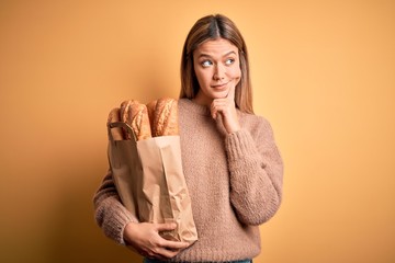 Young beautiful woman holding paper bag with bread over isolated yellow background serious face...