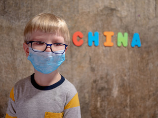 Funny blond kid in big glasses with a green-blue medical mask to protect against influenza viruses in crowded places. China inscription on wall background, Wuhan Coronavirus