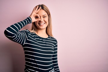 Young beautiful blonde woman wearing casual striped sweater over pink isolated background doing ok gesture with hand smiling, eye looking through fingers with happy face.