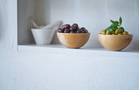 green and brown olives in wooden bowls next to blurred white mortar, atmospheric picture that is suitable for cookbooks