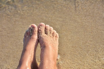 Girls bare feet with golden sand, selective focus. Bare feet in a sea wave.  Relaxation in the ocean.  Woman feet on the sand background. Summertime. Barefoot girl. Feet in the sand