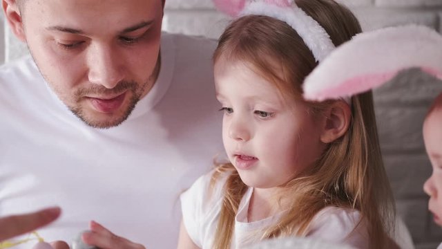 The father and his little daughter are painting the easter eggs into colours with a brush. The daughter is wearing fake bunny ears.