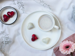 Obraz na płótnie Canvas cup of coffee with cream and excellent bonbons with a heart shape on a white bed. Romantic breakfast in bed. flat lay, top view
