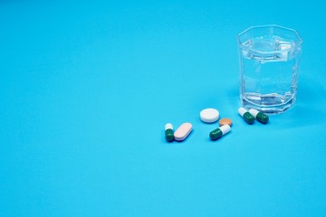 A glass of clear water with pills and capsules for treatment on the blue blckground