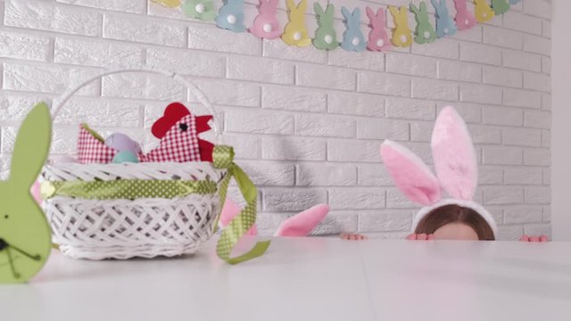 Bunny ears are showing at the table in an easter prepared kitchen. The mother and daughter are wearing bunny ears headbands and having fun on Easter.