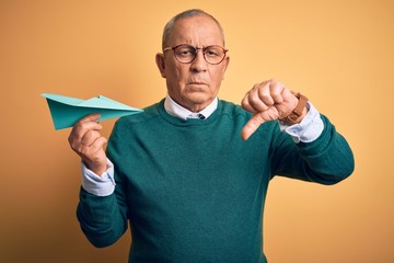 Senior handsome man holding airplane of paper standing over isolated yellow background with angry face, negative sign showing dislike with thumbs down, rejection concept