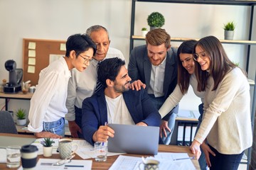 Group of business workers smiling happy and confident. One of them sitting and partners standing around. Working together with smile on face looking at the laptop at the office