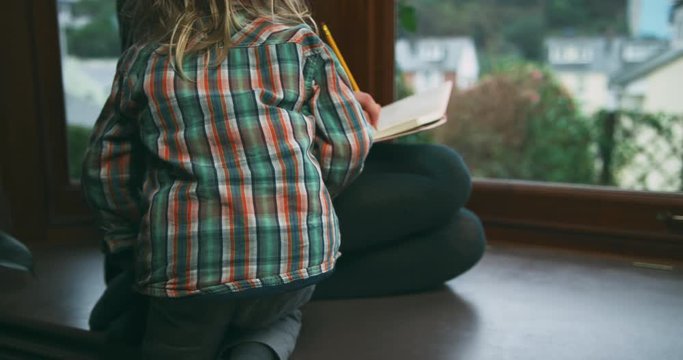 Pregnant woman writing in notebook by window with her little toddler